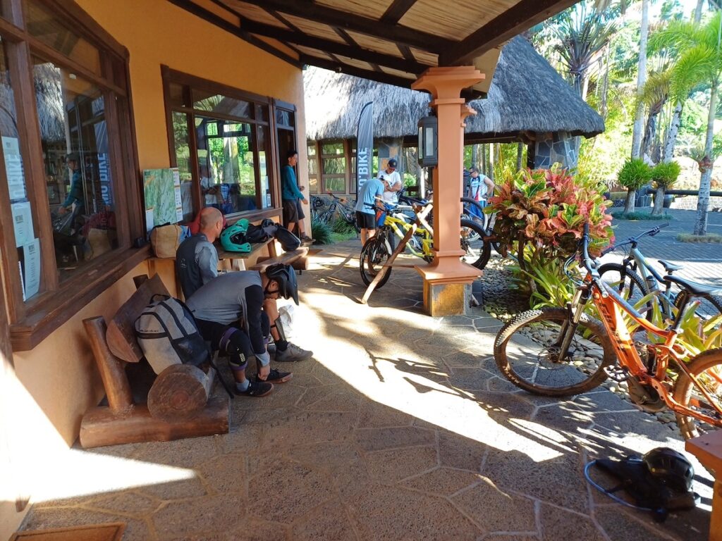 People are preparing for a bike ride outside a building, with mountain bikes parked nearby and some individuals adjusting gear or resting on benches.